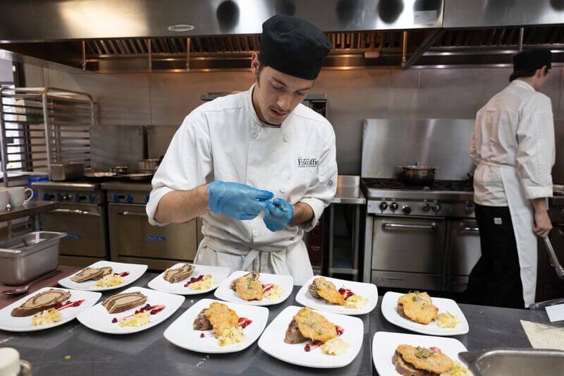 A student chef wearing a white chef's coat and a black skull cap carefully plates several dishes in a teaching kitchen.