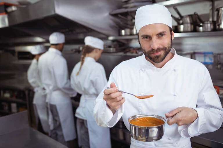 A bearded chef tastes a spoonful of pureed orange food in a professional kitchen