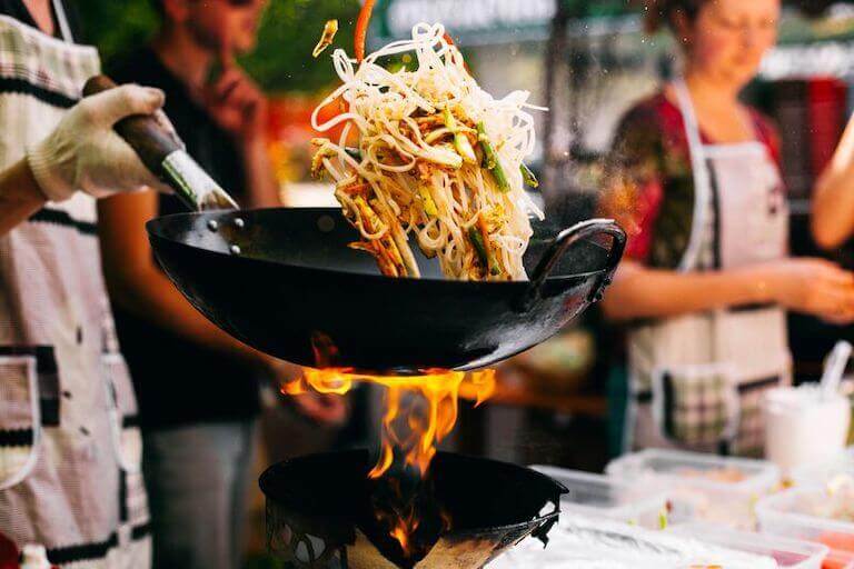 A chef tosses a noodle-based stir-fry using a wok over an open flame.