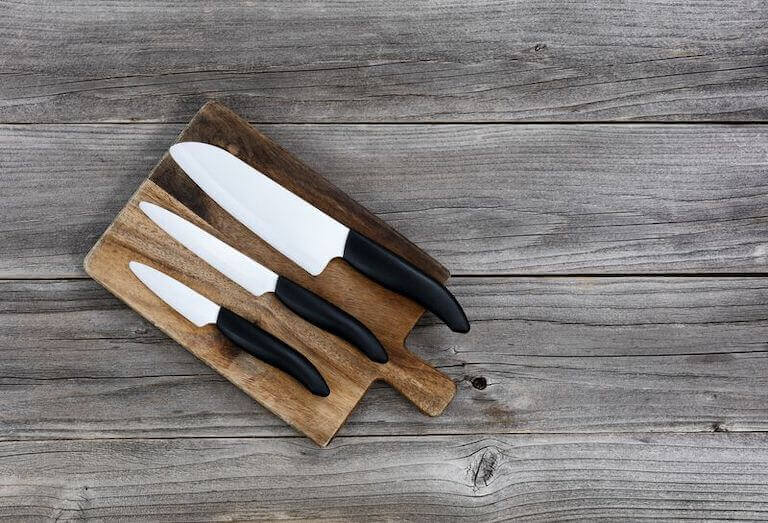 A large, medium, and small knife sit on a wooden cutting board atop a lighter-colored wood working surface.