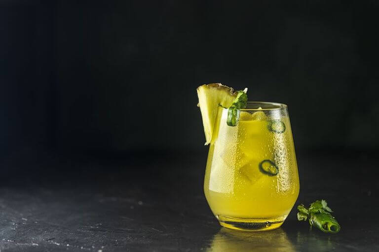 A yellow cocktail with slices of jalapeno and a wedge of pineapple sits on a black countertop