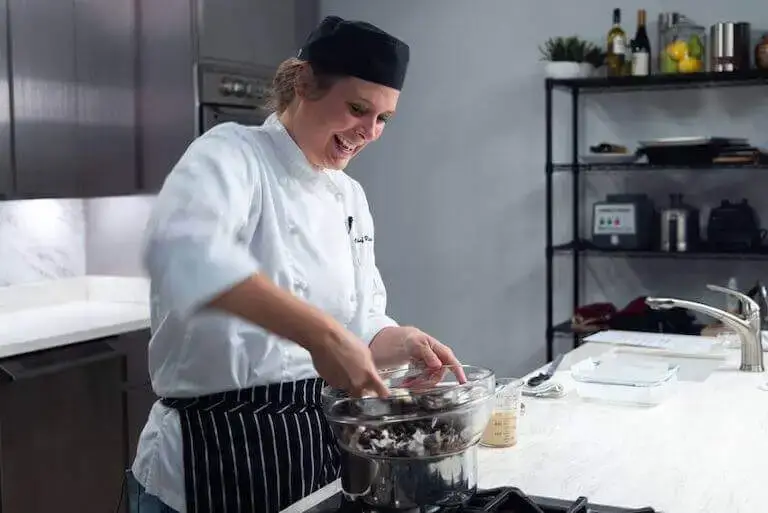 Smiling pastry chef in white coat and black hat stirring chocolate mixture in glass bowl over stove in professional kitchen