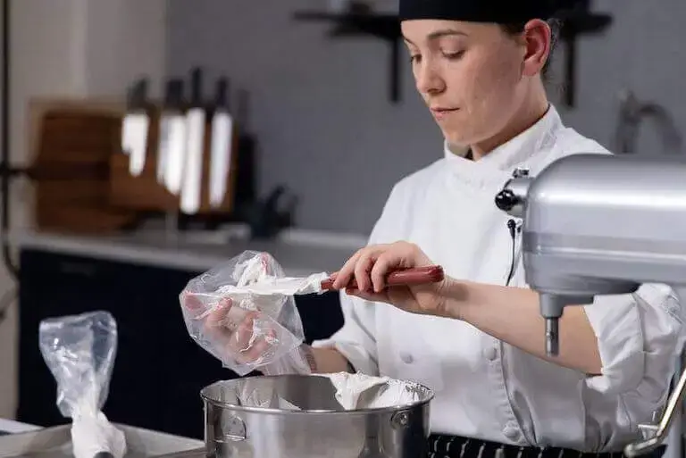 Pastry chef in white coat carefully piping cream onto baking sheet in neat rows using canvas pastry bag