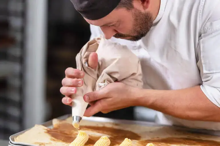 Pastry chef in white coat carefully piping cream onto baking sheet in neat rows using canvas pastry bag