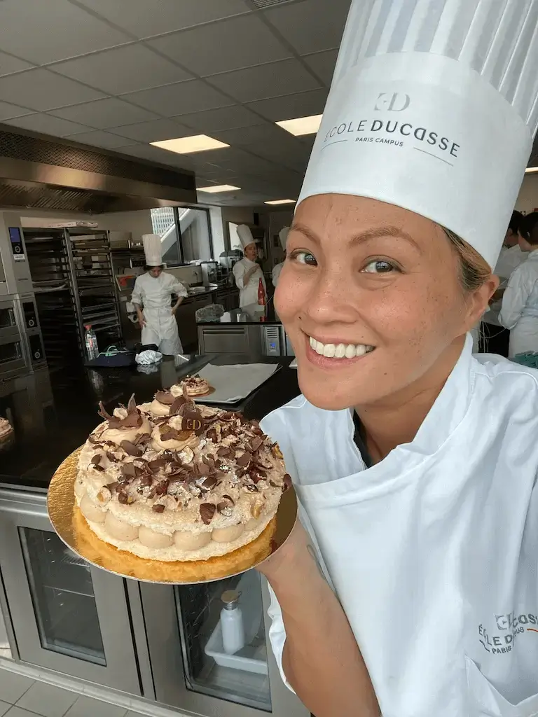 Smiling pastry student in École Ducasse chef's hat holding decorated layered cake in professional training kitchen