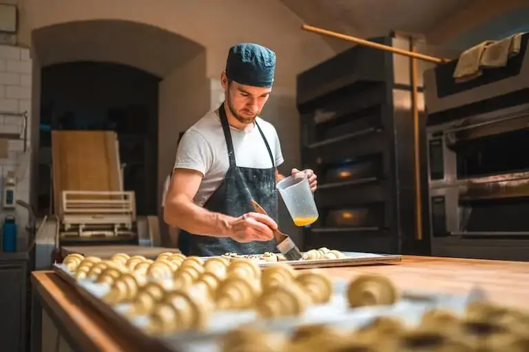 Chef in a commercial bakery brushes egg wash onto a tray of baked goods while holding a measuring cup, with rows of unbaked pastries arranged on a wooden worktable in the foreground.