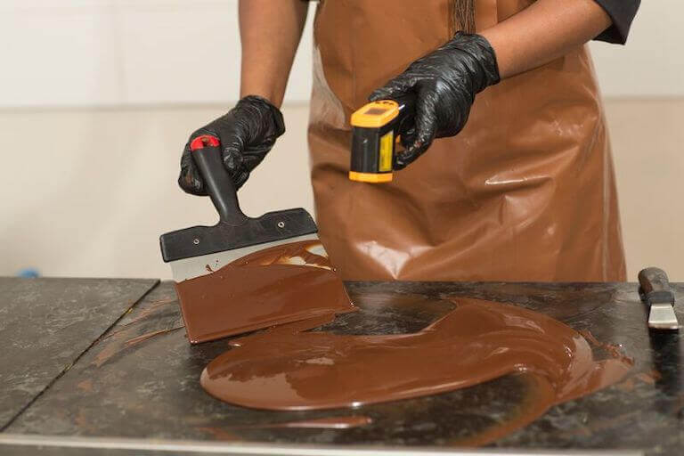 A chef tempers a large quantity of chocolate on a cold marble tabletop, keeping an eye on its temperature with an infrared thermometer.