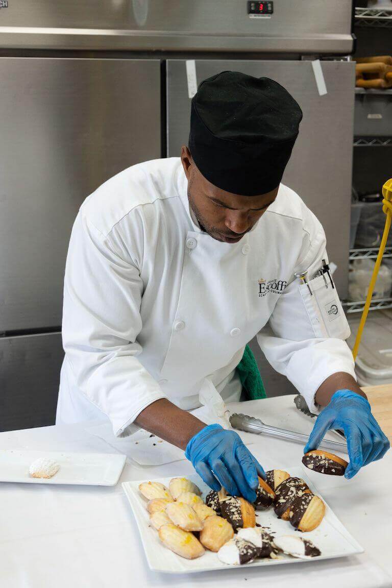 A culinary student in a white uniform plates madeleine cookies in an industrial kitchen setting.