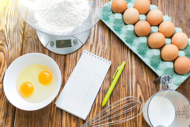 A notepad and pen sit on a wooden countertop surrounded by a carton of eggs, measuring cup, whisk, digital scale with flour in the bowl, and a ceramic bowl with two eggs cracked in it.