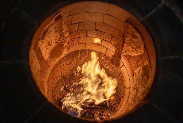 Looking down into the interior of a traditional Turkish wood-fired stone brick oven, with flames casting an orange glow and naan stuck to its sides.