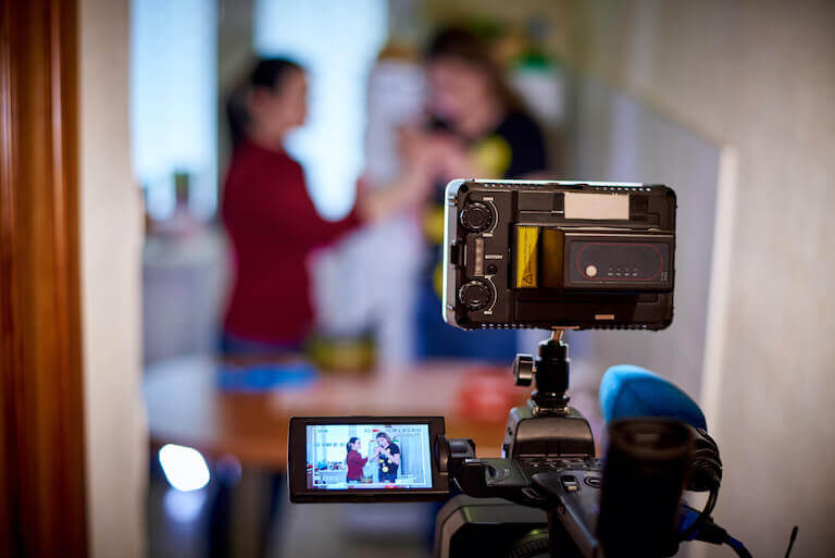 The back screen of a professional camera displaying two people recording a home cooking segment in a small kitchen.