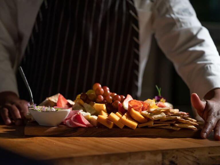 A charcuterie board of assorted cheeses, grapes, crackers, meat and a dish of spread rests on a table framed by the chef’s hands as it is set down.