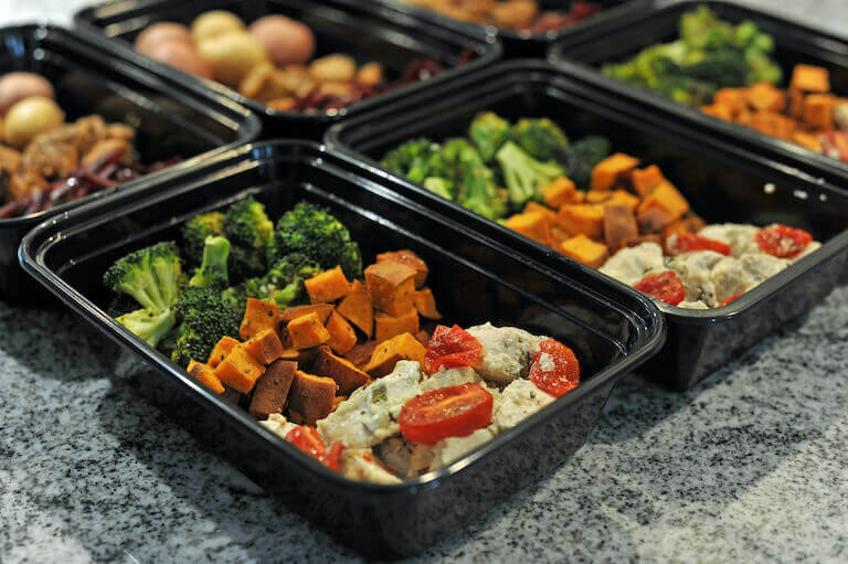 A collection of black plastic containers containing prepared meal components, including cooked broccoli, sweet potatoes, and chicken salad with cherry tomatoes.
