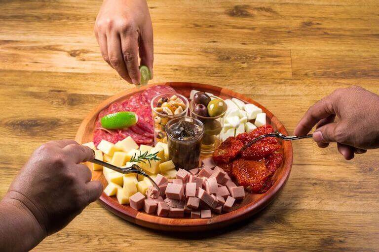 A compilation of ham, pepperoni, sun-dried tomatoes, and cheeses circle cups of pimento olives, nuts, and sauce on a circular wooden board as hands reach in to serve the food.