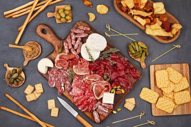 A dark wooden charcuterie board containing a dense collection of smoked meats alongside three wheels of white soft cheese and surrounded by various tools, crackers, and breadsticks.