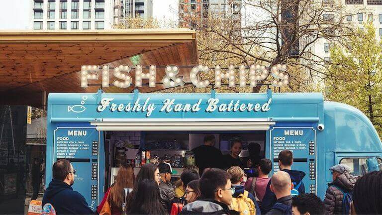 A large crowd congregates in front of a blue food truck advertising fish and chips in an urban setting.