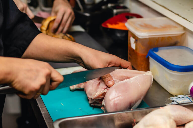 Close-up of hands butchering a raw chicken on a green cutting board in a restaurant kitchen, with plastic containers of other prepped ingredients nearby.