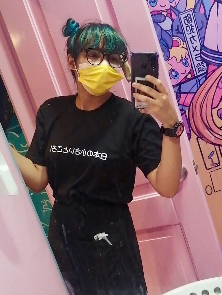 Estefania Colamarco takes a selfie in the bathroom mirror of a restaurant. She is wearing a face mask, her hair is dyed blue, and she wears a black server’s apron and black tee shirt.