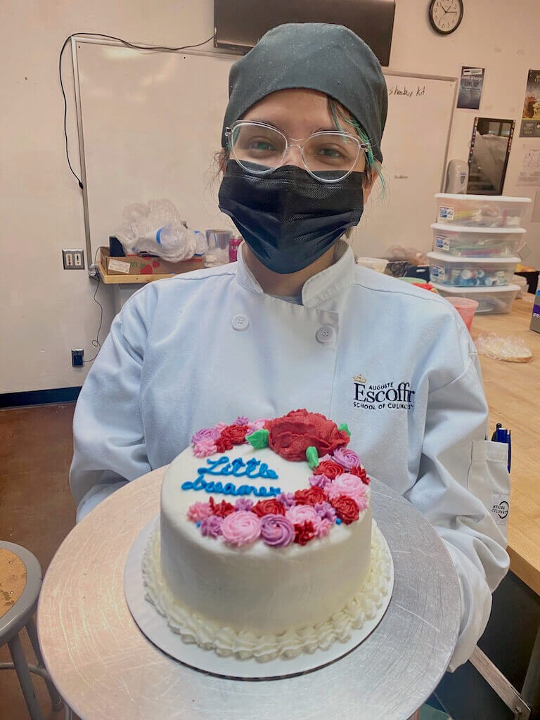 Estefania Colamarco wears a face mask and holds a cake she made and decorated while a student at Escoffier