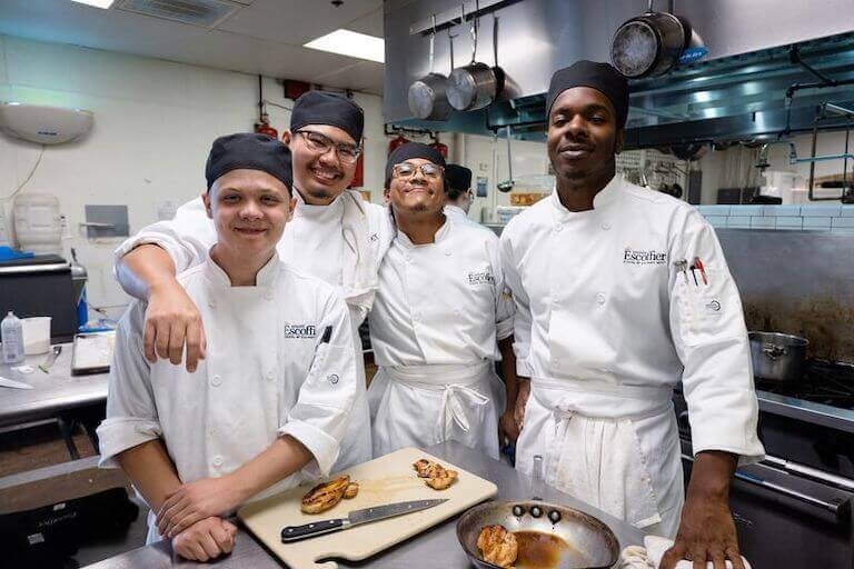 Four Escoffier classmates in matching white uniforms stand smiling for a group photo in a professional kitchen.
