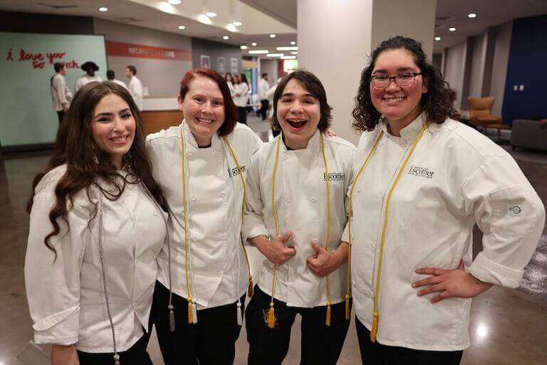 Four culinary students, all smiling, wearing graduation attire at culinary school.