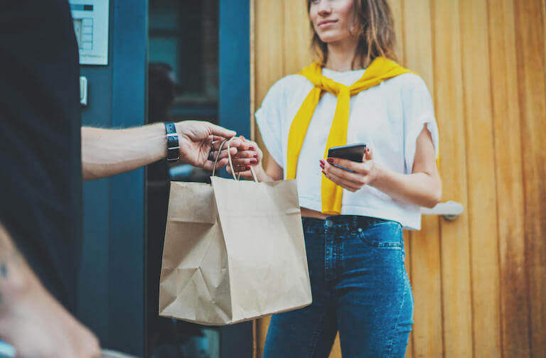 Male courier handing a brown paper delivery bag to a female customer who is standing outside of a door and holding a cell phone.