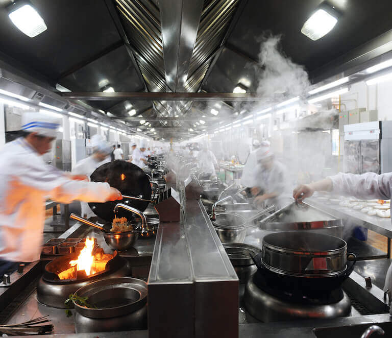 Many cooks work side-by-side in a busy restaurant kitchen, stirring pots, prepping ingredients, and performing other tasks as fire and steam rise from their work stations.