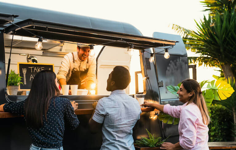 Three customers stand at the window of a food truck chatting with the owner in a tropical setting.