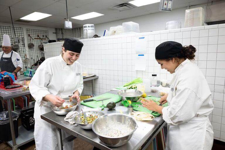 Two culinary students smile while mixing diced vegetables in silver bowls in a professional kitchen.