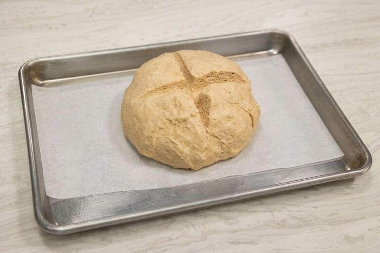 A round ball of dough with a deep x carved into its top, sitting on parchment paper on a baking sheet.