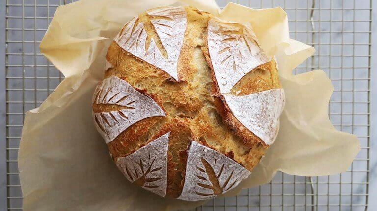 A loaf of sourdough bread with decorative leaf scoring sits on a metal cooling rack on a marble countertop.