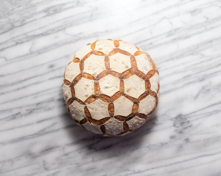 Overhead view of a loaf of artisan bread on a marble cutting board, with bread scored in a hexagonal pattern. 