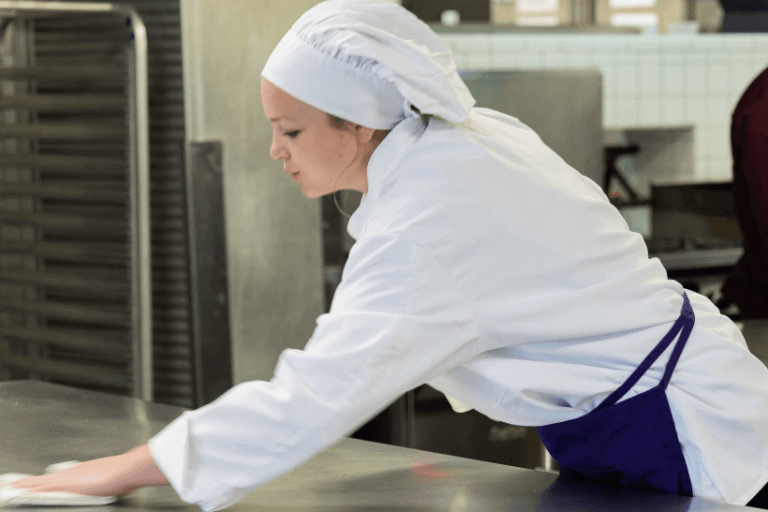 A cook wearing a white uniform in a professional kitchen cleans the stainless steel surface of their workstation.