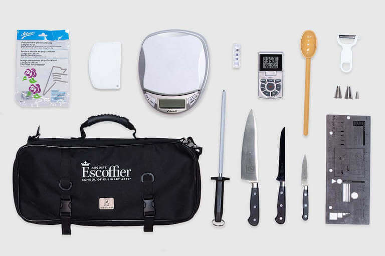 An overhead view of the items included in an online student's toolkit. Some of the items include a German steel sharpener, a polyurethane pastry bag, paring knife, boning knife, 8-inch chef's knife, kitchen scale, a hi-temp spoon, and other materials.