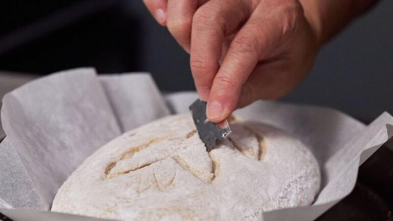 Hand holding a razor blade adding decorative scoring to a loaf of sourdough bread.