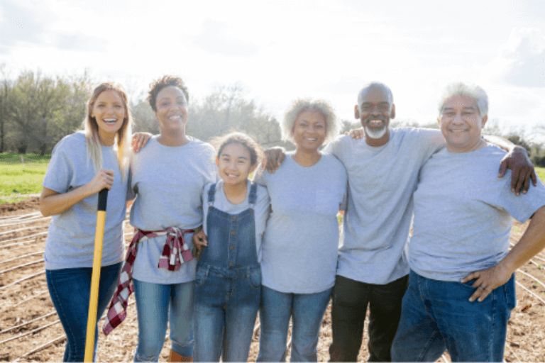 Several people of different ages and ethnicities wearing t-shirts and jeans stand in a wide open field with their arms around one another and smiling.