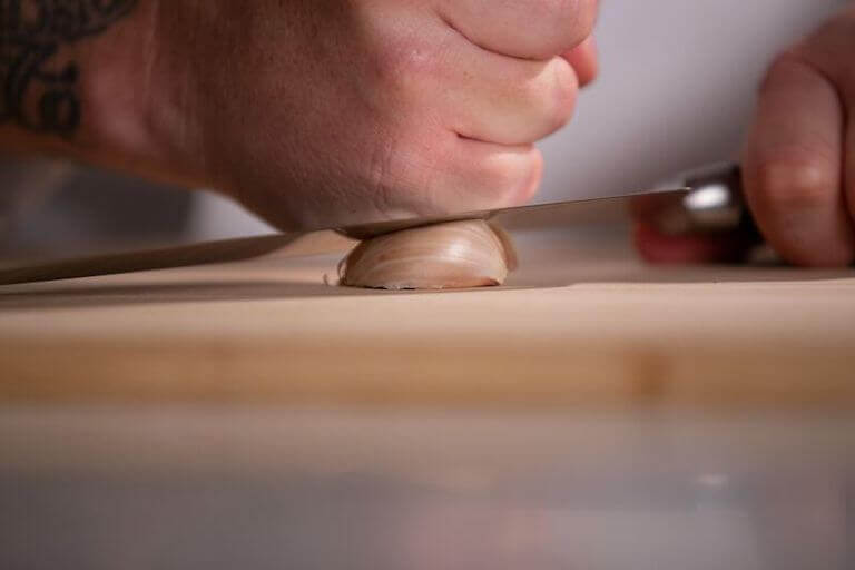 Two hands use the side of a chef’s knife to press a clove of garlic into a wooden cutting board. 