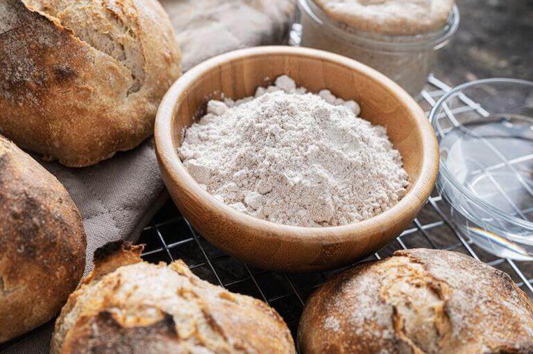 Wooden bowl of whole wheat flour in the middle of freshly baked sourdough bread buns.