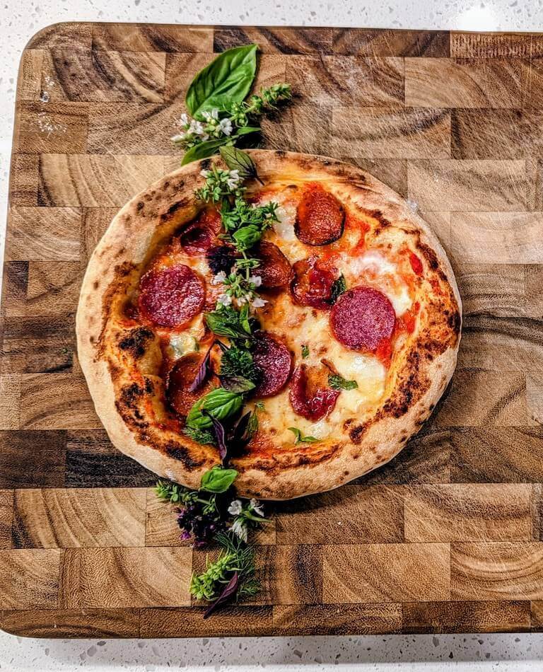 A handmade pepperoni pizza on a wooden cutting board, garnished with whole basil leaves and flowers.