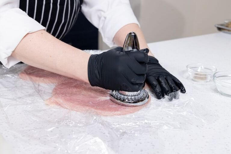 A pair of hands in black gloves, holding a meat tenderizer over a large piece of turkey that's been pounded flat and is covered in plastic wrap.