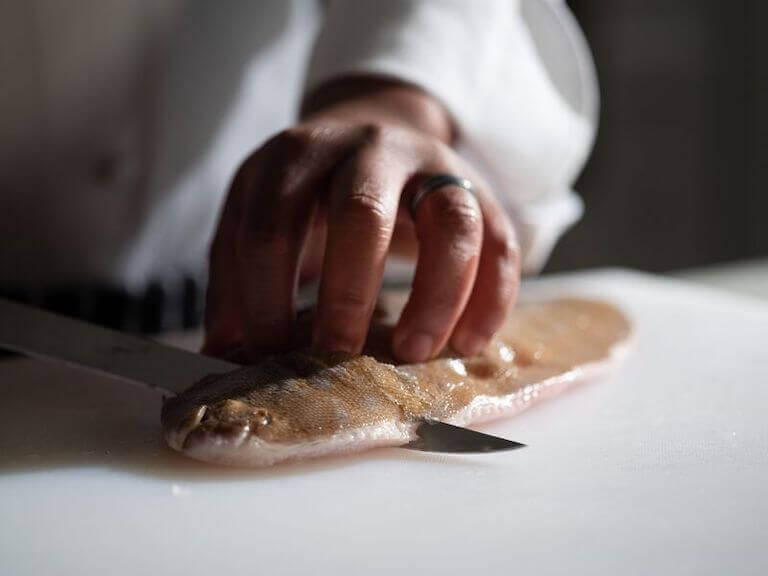 Person’s hand holding down round fish while using a knife to fillet.