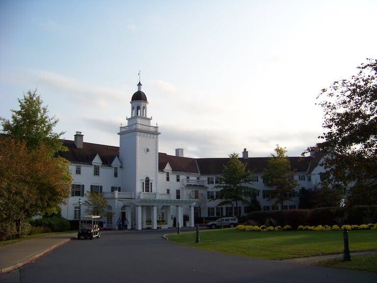 The white, colonial-style facade of a grand hotel, with a tall tower and large columns, overlooks a wide driveway and a landscaped garden.