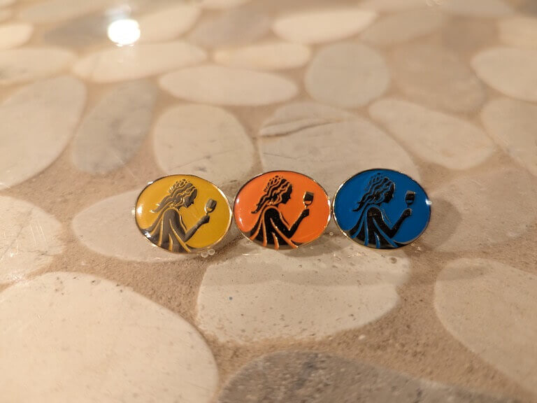 Three colorful pins, each depicting an image of a woman examining a glass of wine, sit side by side on a stone countertop.