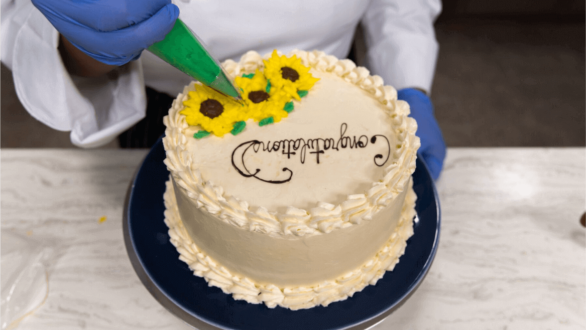An Escoffier pastry student pipes frosting sunflowers and leaves on a vanilla cake
