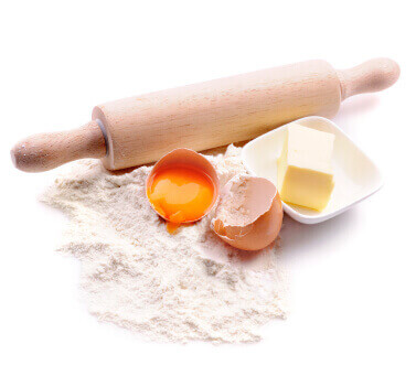 Flat view of baking ingredients: flour, eggs, butter, and a rolling pin on a white table