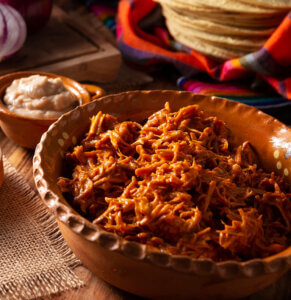 A bowl of cochinita pibil with a stack of corn tortillas.