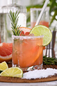 Paloma cocktail in a rocks glass, with salt on the rim and garnished with a lime wheel, grapefruit wedge, and rosemary sprig.