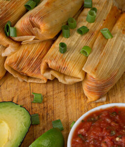 Tamales on a wood cutting boar with a sliced avocado and bowl of salsa
