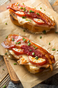 Two open-faced sandwiches on a wooden table, each with a toasted baguette slice topped with melted cheese, tomato, crispy bacon, and chives.