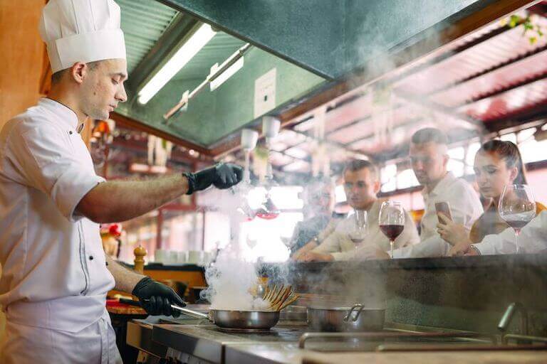 A chef dressed in a traditional chef’s uniform cooks behind the counter, where customers watch as they drink wine.